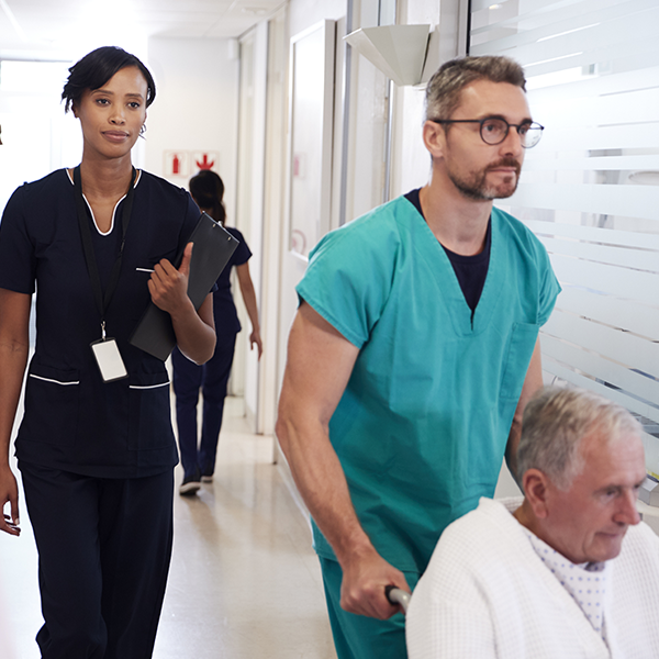 Male nurse pushing elderly male patient in wheelchair in busy hospital hallway