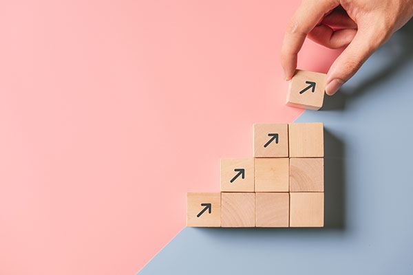 Pink and blue background with wooden building blocks set upon background in a stepped up formation, hand adding final block