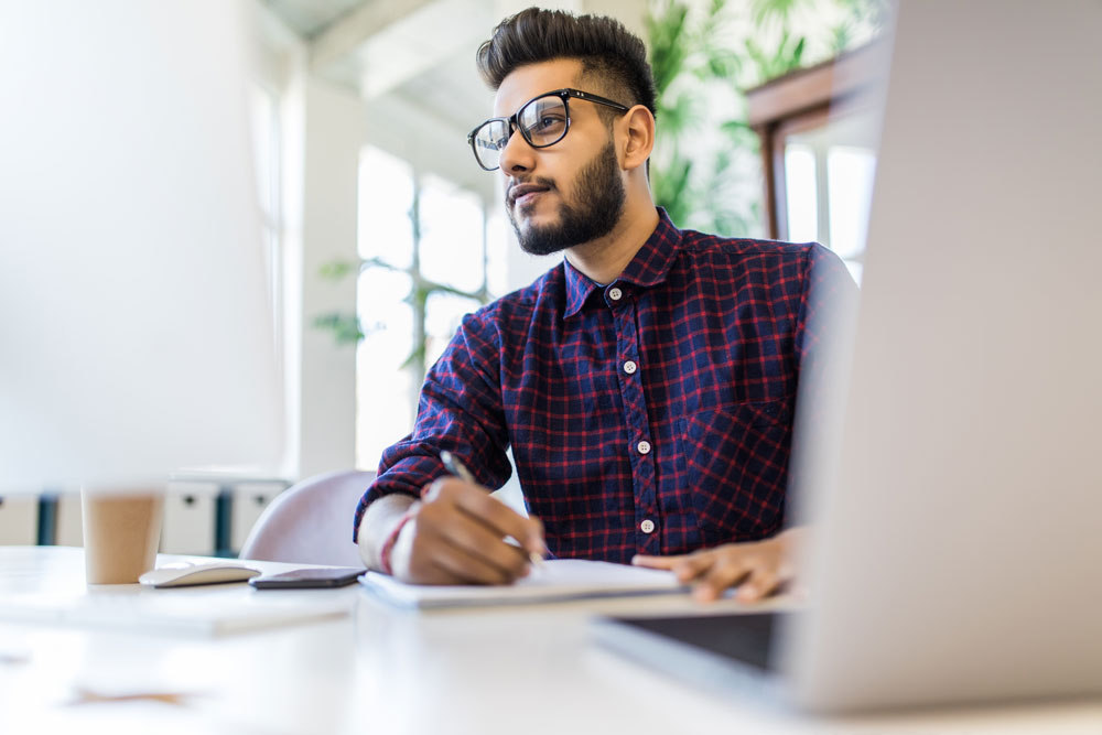 Young businessman wearing buttoned up shirt, writing on paper, looking at computer