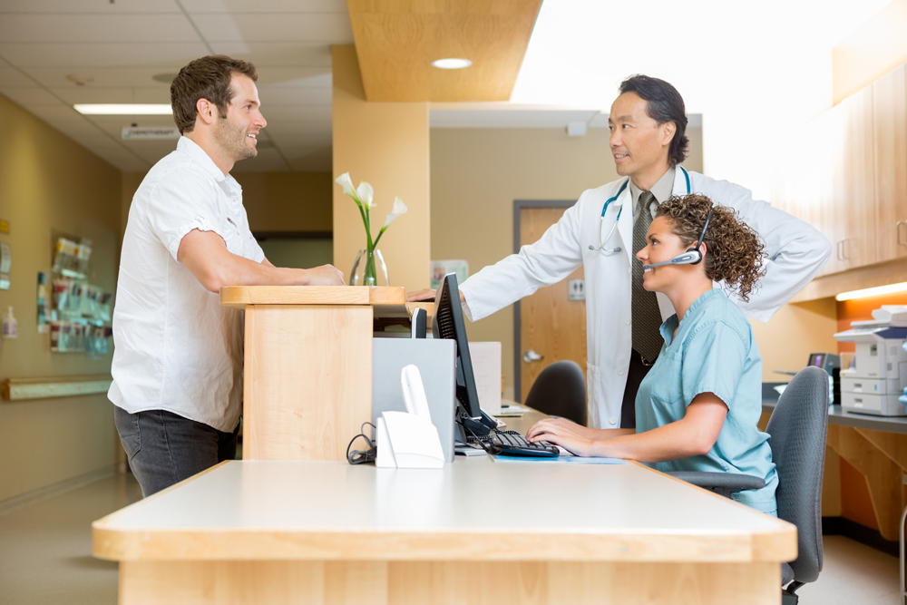 Male patient standing at medical office front desk, male doctor and female nurse on other side