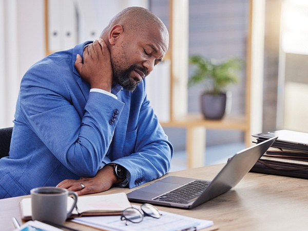 Man in office setting rubs neck in pain as he sits in front of his laptop.